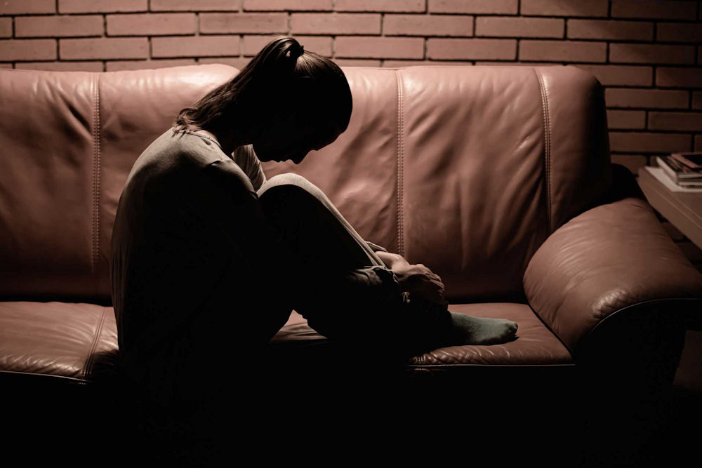 Silhouetted individual sitting alone on a couch in a dark room, representing isolation and emotional distress linked to symptoms of untreated depression.